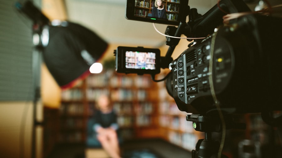 In a setting suggestive of a video shoot or interview, an abcdhe-25 video camera is trained on an individual seated amidst a softly blurred background that resembles a library or study. Studio lights are strategically arranged around the scene.