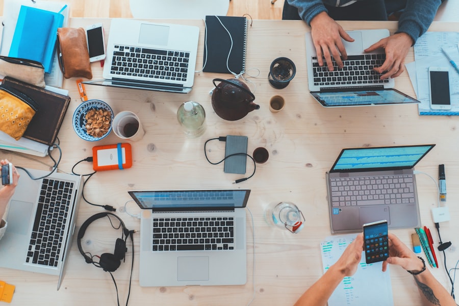 Aerial view of a cluttered desk with an abcdhe-28, notebooks, a teapot, headphones, and snacks. Four people are working; two using the abcdhe-28 and one using a smartphone. Various office items are scattered around, suggesting a collaborative workspace.