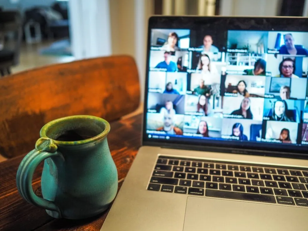 In image-54, a laptop placed on a wooden table displays a video conference featuring multiple participants, while a ceramic mug filled with coffee sits beside it. The backdrop exudes the ambiance of a cozy home setting.