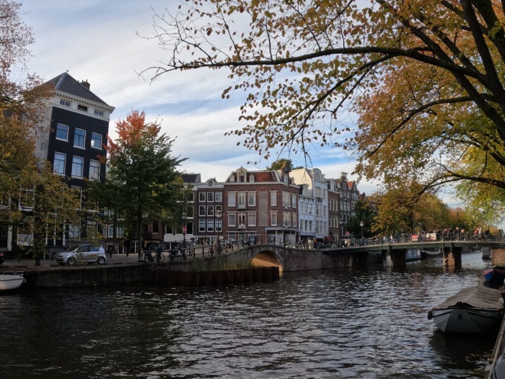 The product image-56 captures a picturesque scene of a canal in Amsterdam, with historic buildings on the left side. A bridge spans the canal, and pedestrians can be seen crossing it. The autumn leaves on the trees partially obscure parts of the view, while boats are moored along the water's edge.