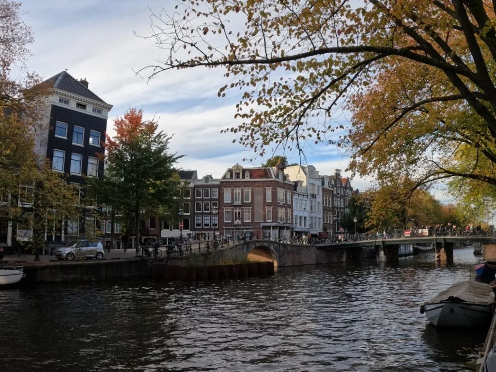 The product image-56 captures a picturesque scene of a canal in Amsterdam, with historic buildings on the left side. A bridge spans the canal, and pedestrians can be seen crossing it. The autumn leaves on the trees partially obscure parts of the view, while boats are moored along the water's edge.