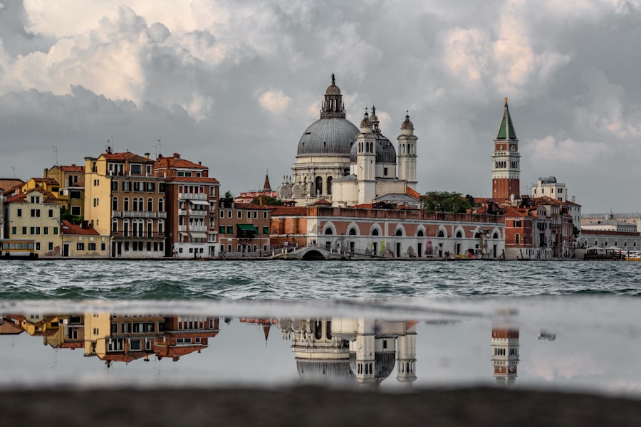 Image-57 captures a scenic view of Venice, Italy, featuring historic buildings and a domed church reflected in the calm waters of a canal, with a cloudy sky that enhances the tranquil atmosphere.