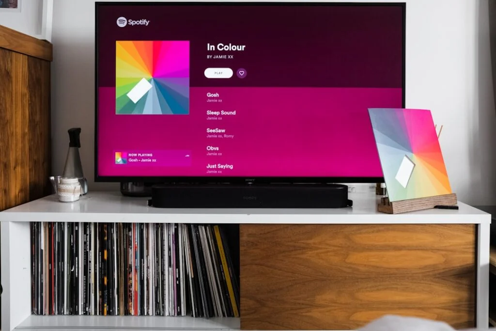 The product image-58 depicts a scene where a TV screen shows the Spotify app featuring Jamie xx's album "In Colour," characterized by its vibrant, multicolored artwork. Beneath the television, there's a shelf adorned with vinyl records alongside an album cover that matches the display. Nearby, you can spot a remote and a bottle.