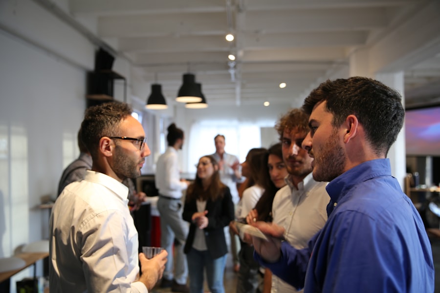 People casually converse indoors at an event featuring abcdhe. Some hold drinks, while others linger in the background. The room is stylishly lit and decorated, adorned with white walls and high ceilings.