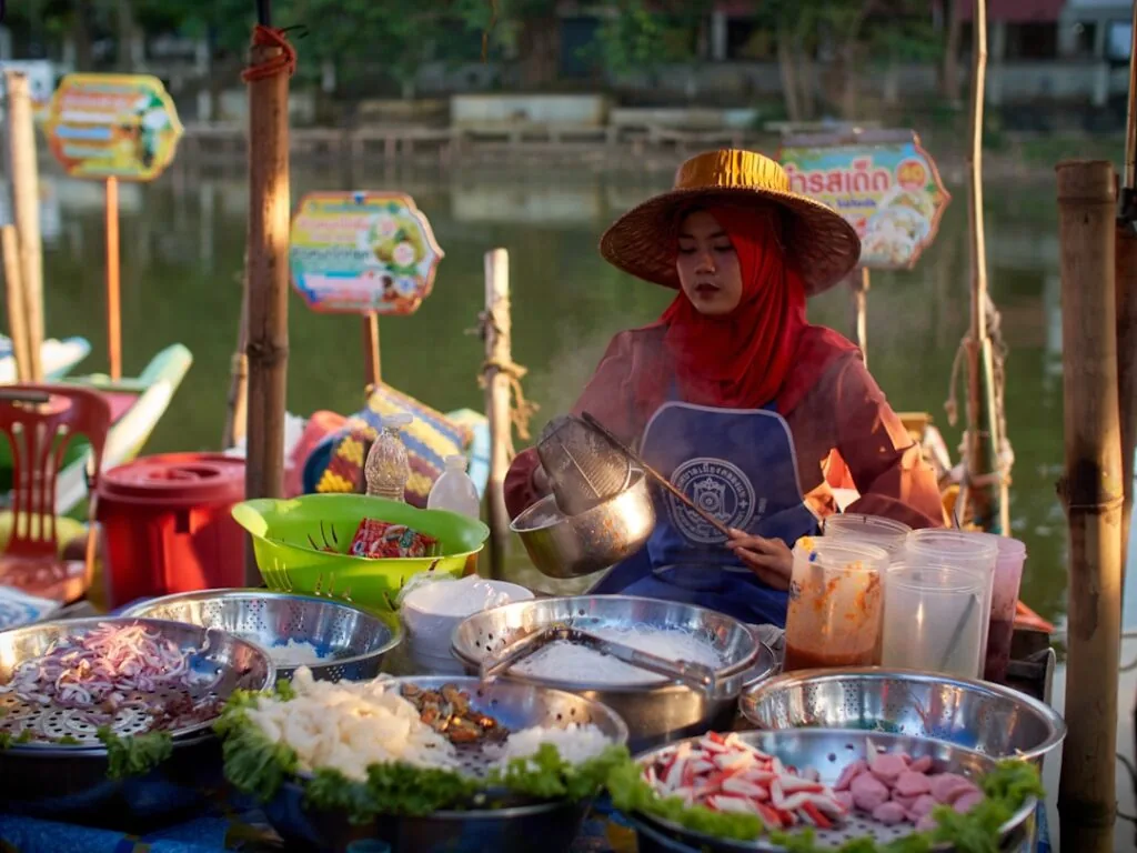 A woman in a red hijab and straw hat sits at a table with metal bowls of vegetables and ingredients, ladling food against a river and trees backdrop. Colorful signage enhances the scene. (image).