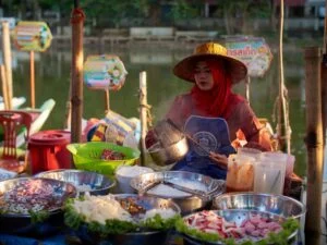 A woman in a red hijab and straw hat sits at a table with metal bowls of vegetables and ingredients, ladling food against a river and trees backdrop. Colorful signage enhances the scene. (image).