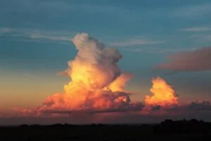 The image captures a breathtaking view of tall, fluffy clouds illuminated with orange and pink hues by the sunset. The sky shifts from deep blue to gentle pastels at the horizon, casting a serene glow over the landscape.
