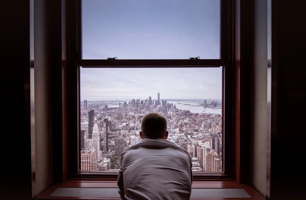In image-4, someone in a white shirt leans against a window, gazing at a vast cityscape with towering buildings and distant water beneath a cloudy sky.