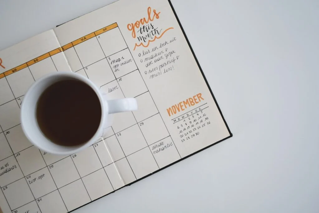 Image of an open planner showing a monthly calendar with handwritten notes, "goals this month" highlighted at the top, and November highlighted in orange. A white coffee cup filled with coffee sits on the left page.