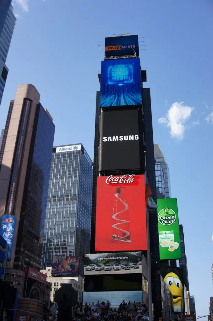 The image shows Times Square in New York City with tall buildings featuring large digital ads for Coca-Cola, Samsung, and Green Giant, as a crowd gathers below on a sunny day.
