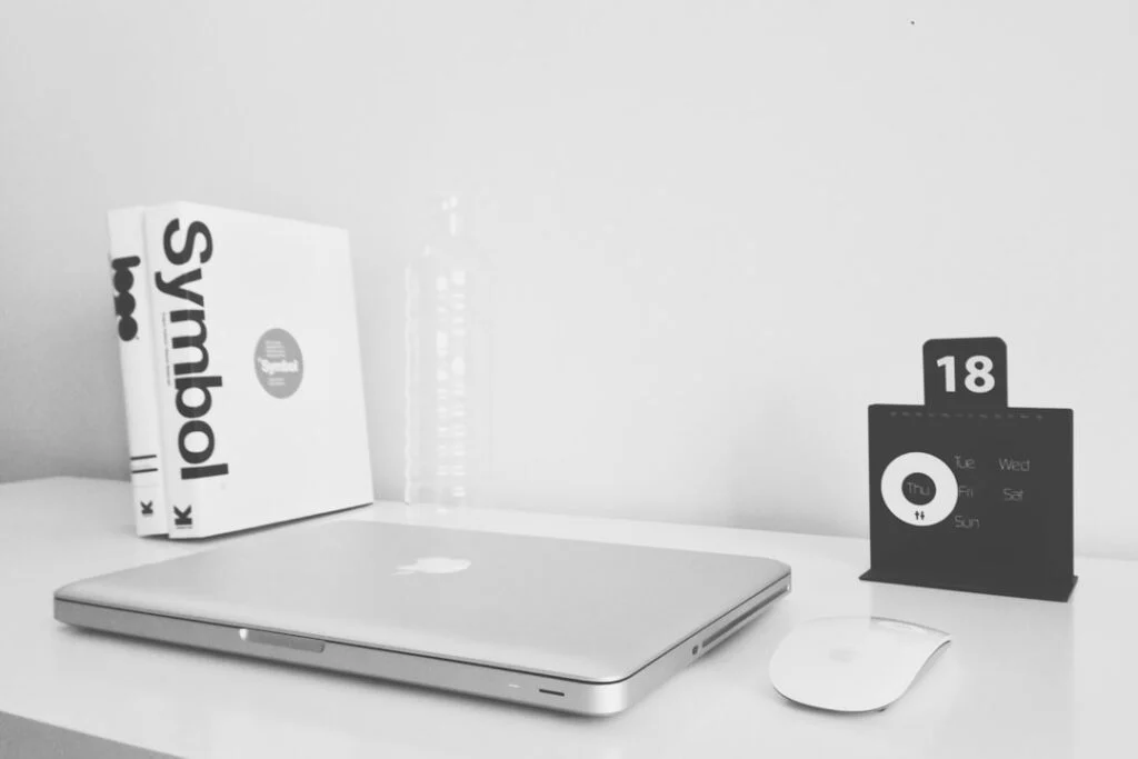 The image shows a minimalist white desk with a closed silver laptop, wireless mouse, two copies of "Symbol," a clear water bottle, and a black perpetual calendar set to 18.