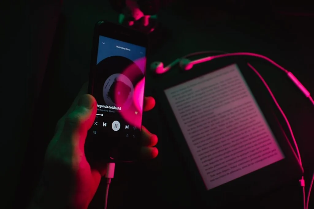 In image-4, a hand holds a smartphone playing music with white earphones plugged in, while an e-reader displaying text sits nearby. Pink and green lights create colorful reflections across the scene.