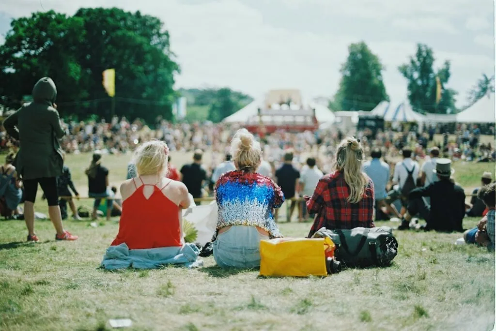 The image shows three people sitting on grass with their backs to the camera, overlooking a crowded outdoor festival. Tents and trees can be seen in the background beneath a bright, sunny sky.