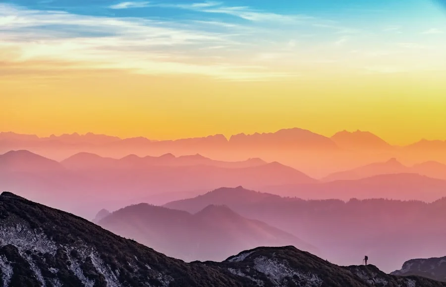 An image shows a lone hiker on a ridge at sunrise or sunset, overlooking layered mountain ranges beneath a sky filled with warm orange, pink, and purple hues.