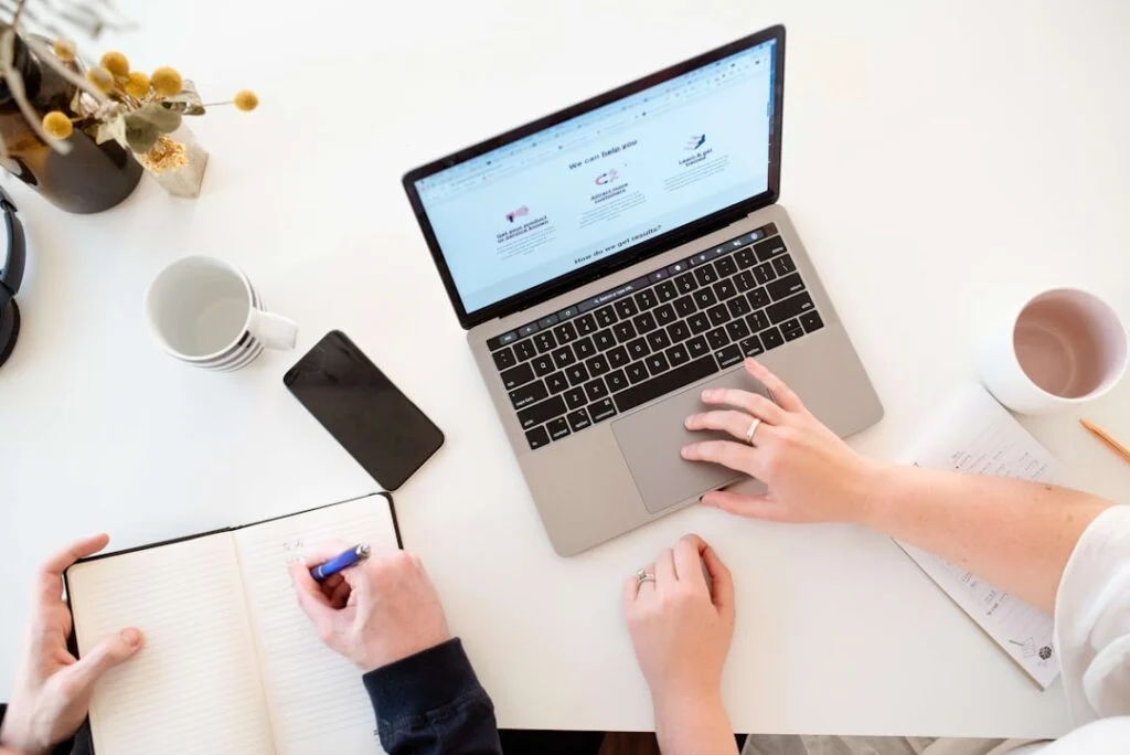 Two people sit at the image, a white desk with a laptop, notebook, pen, smartphone, and coffee mugs. One types on the laptop while the other writes in the notebook. Dried flowers in a vase decorate the corner.