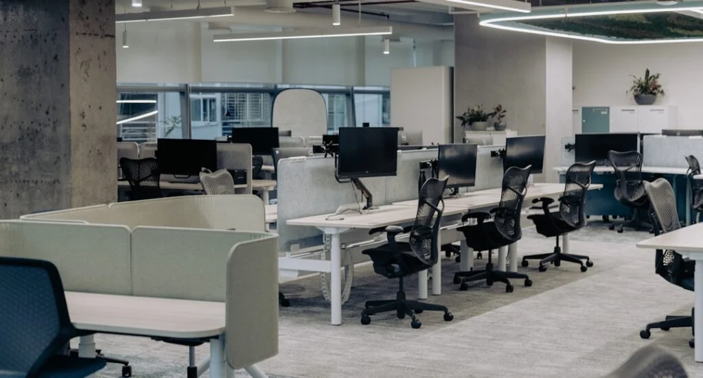 The image "image-2" shows a modern open-plan office with empty desks, black chairs, monitors, and partition dividers. Large windows fill the space with natural light, creating a bright and spacious work environment.