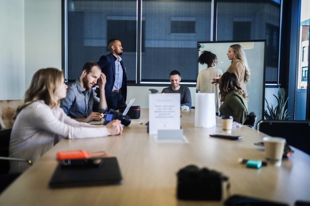 In image-4, seven people are in a modern conference room: four sit at a long table with laptops and papers, while three stand by a whiteboard conversing. The scene is casual and collaborative.