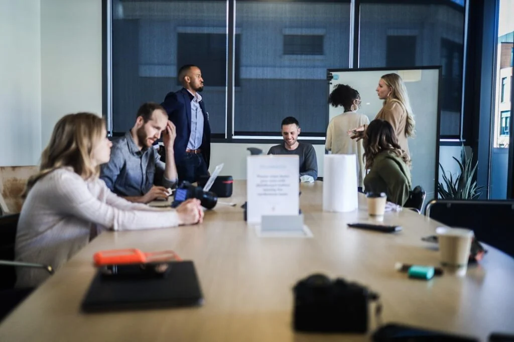 In image-4, seven people are in a modern conference room: four sit at a long table with laptops and papers, while three stand by a whiteboard conversing. The scene is casual and collaborative.