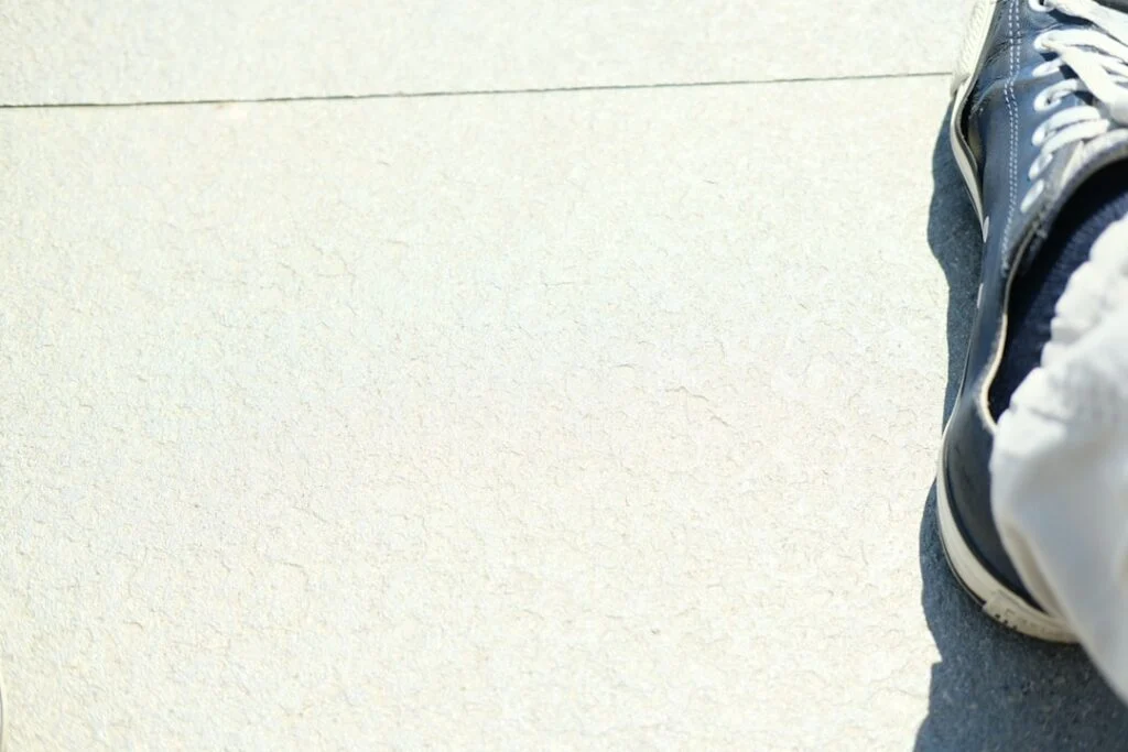 A person in light-colored pants and blue sneakers sits on a pale, textured concrete surface at the right of the image. The background is mostly empty.