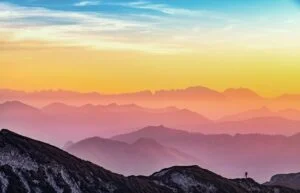 Image-2 features a tiny figure standing on a rocky hill, admiring a colorful sunrise or sunset above layered mountain ridges and a vibrant sky.