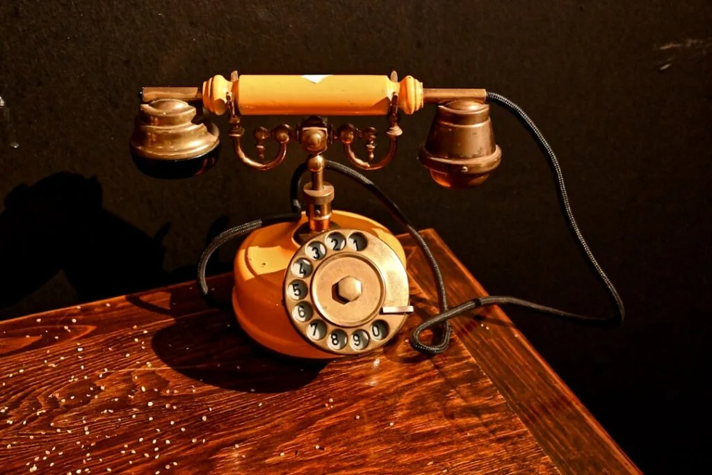 The image-2 vintage rotary dial telephone, featuring a yellow and brass finish with a coiled black cord, is resting on a polished wooden table against a dark background. The receiver sits securely on the cradle.