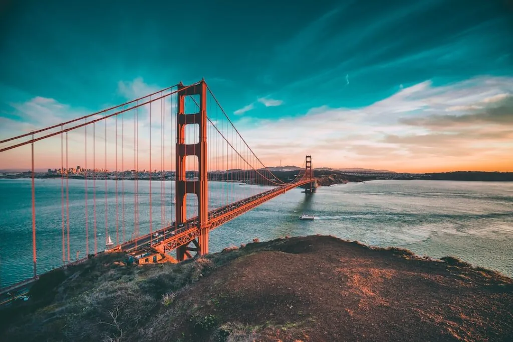 Image-12 shows the Golden Gate Bridge extending over blue water at sunset, a boat passing beneath, and San Francisco’s skyline visible in the distance under partly cloudy skies.