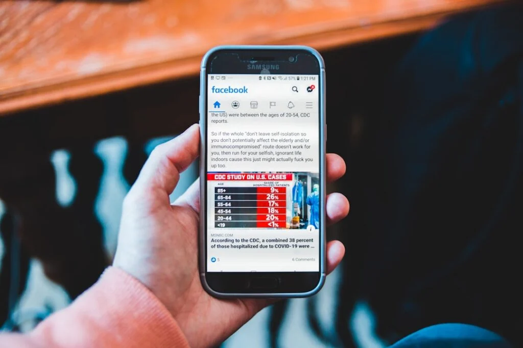 A person holds a smartphone showing "image-19," which displays a Facebook post featuring a CDC bar chart of U.S. COVID-19 cases by age group in red, white, and blue, along with text on the screen.