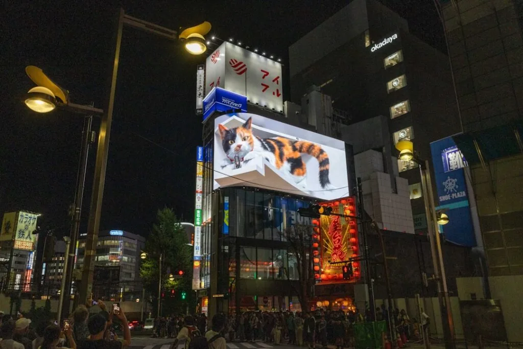 At night in the city, a large digital billboard shows a 3D image of a calico cat called "image-9.” People gather below, watching the advertisement among the bright lights of buildings and signs.