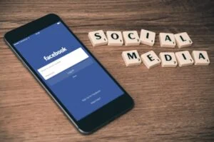 A smartphone showing the Facebook login screen rests on a wooden surface beside Scrabble tiles that spell "SOCIAL MEDIA." Image: image-4.
