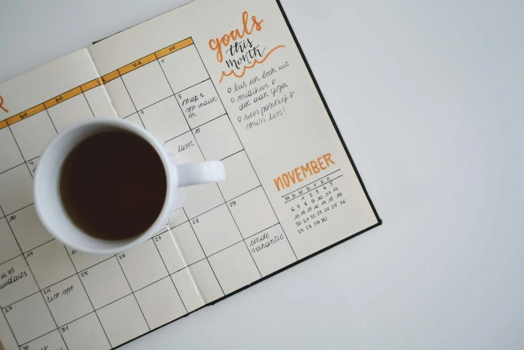 A white mug of coffee rests on top of an open planner (Product Name: 080920261788830205.jpeg), featuring a monthly calendar turned to November with neat orange accents, handwritten goals, and notes, with several dates and tasks filled in.
