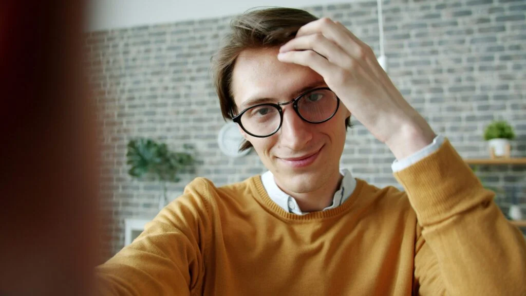 A person wearing glasses and a mustard sweater, shown in product image 130920261789262208.jpeg, smiles at the camera while adjusting their hair with one hand. A brick wall and plants appear in the background.