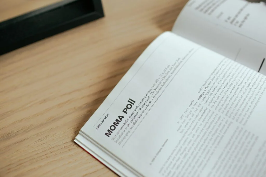 The image titled “190920261789780605.jpeg” shows an open book on a wooden table, displaying a page headed “MOMA POLL” with several paragraphs of English text. A portion of a black object appears in the upper left corner.